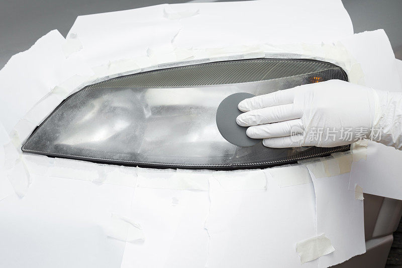 修理工手戴白色橡胶防护手套，用砂纸打磨汽车大灯。改造的过程。恢复汽车灯。特写镜头。前视图。