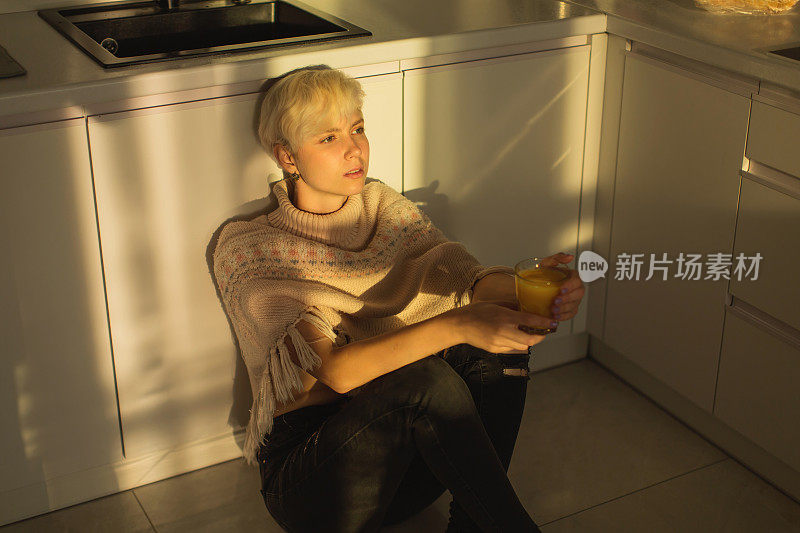 金发女人端着一杯橙汁坐在厨房的地板上。