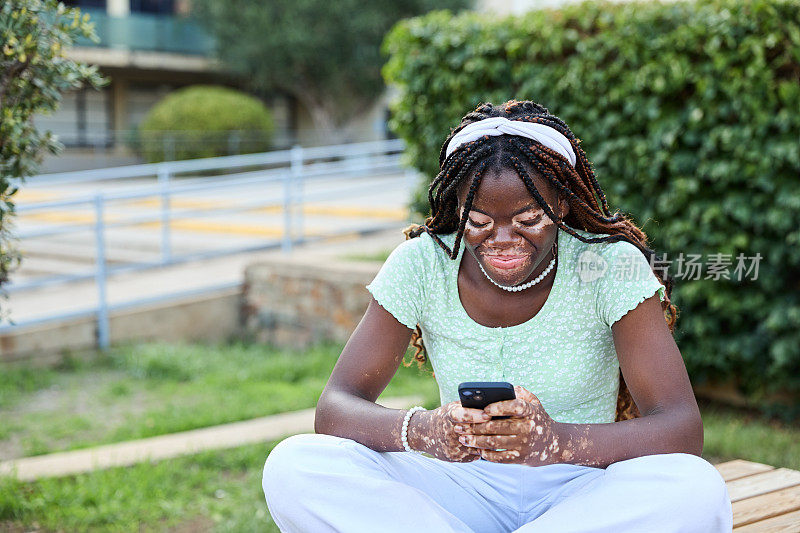 患有白癜风的年轻黑人女学生坐在大学外的长凳上玩手机。青少年文化与科技理念。手机和社交媒体成瘾