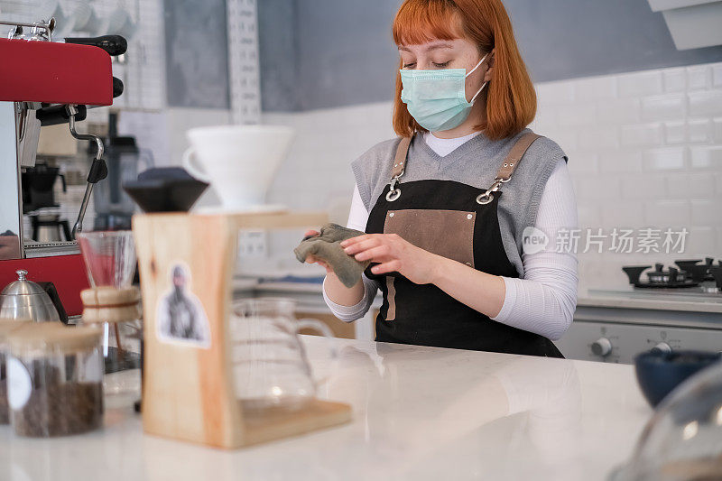 年轻的女咖啡师戴着口罩和清洁咖啡店吧台
