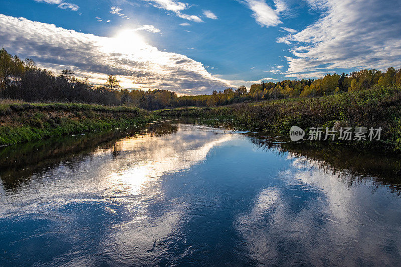 天空与云彩和太阳的倒影在一条小河的蓝色水面上。