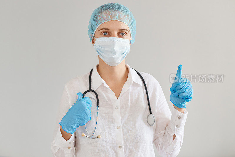 室内拍摄阳性女医生身穿医疗制服，戴着手术口罩，站着，看着镜头，翘起大拇指，批准了新的病毒治疗方法。