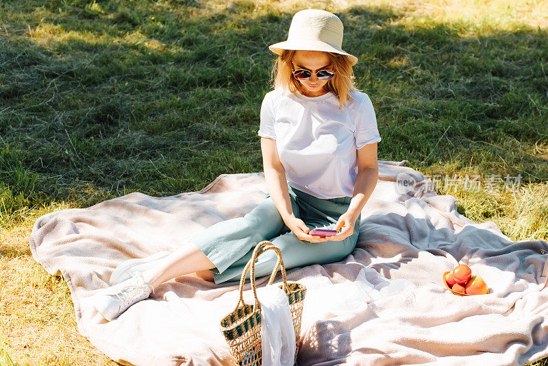 美丽的年轻女人戴着太阳镜和帽子在大自然中使用手机度假。微笑的女孩看着智能手机和放松毯子上的绿色草坪，周末野餐在夏天