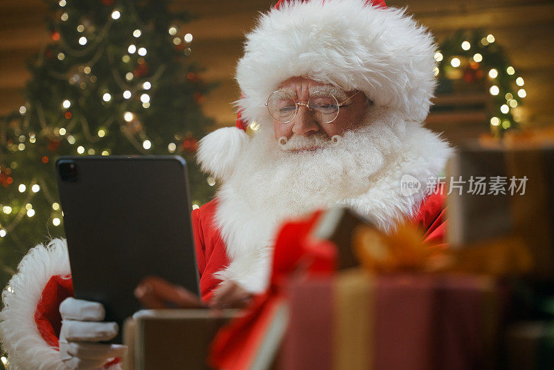 圣诞老人坐在装饰着圣诞彩灯和圣诞树的办公室里，正全神贯注地看着手中的数码平板电脑，前景是礼物盒