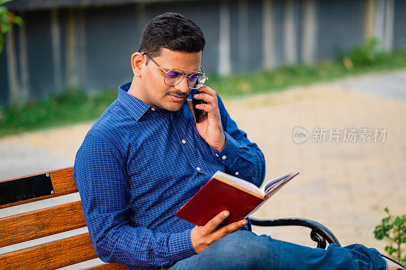 一个年轻的学生坐在长凳上读笔记本和讲电话。