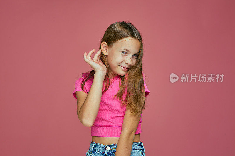 微笑，尴尬，害羞的白人女孩固定她的头发在粉红色的工作室背景。孩子们的时装