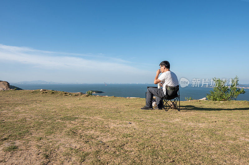 在野外的海边，一名亚洲男子坐在简单的桌椅上喝水