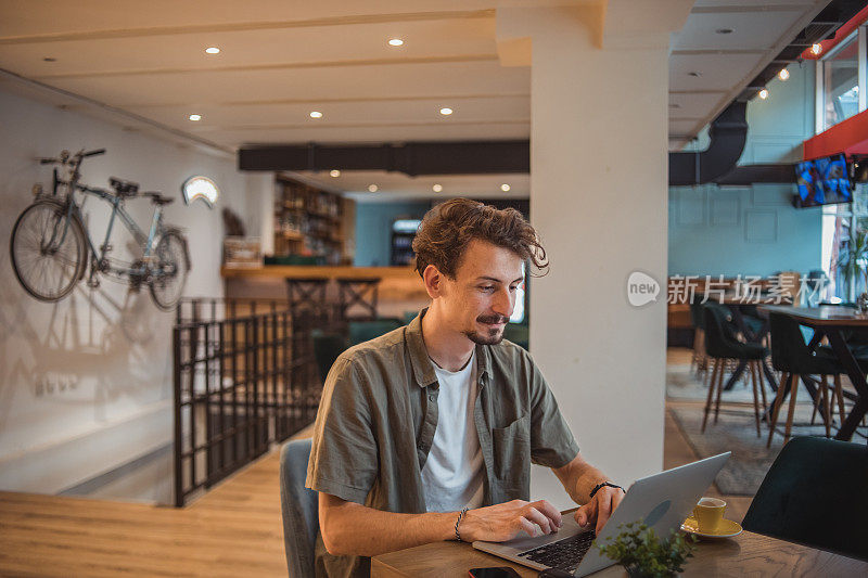 一位年轻的自由职业者坐在咖啡店里，一边喝着桌上的浓缩咖啡，一边在笔记本电脑上工作