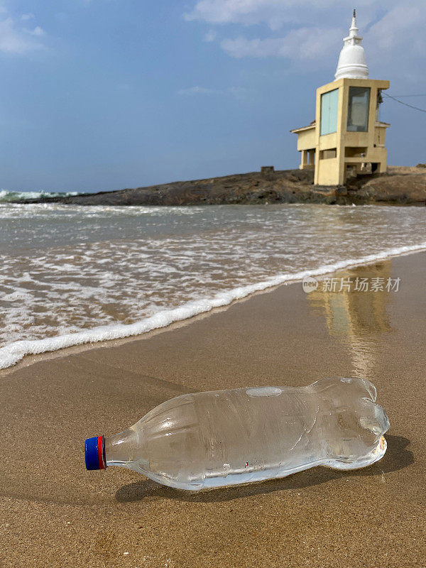 特写图片，空的，透明的塑料饮料瓶，蓝色盖子被冲到低潮水的沙滩上，海浪把海洋垃圾和污染冲到岸边，乱扔的沙子，肮脏的海滩，大海和岩石背景