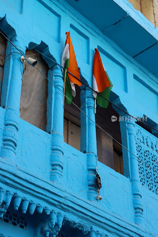 印度拉贾斯坦邦焦特布尔蓝色建筑外墙阳台上飘扬的两面印度国旗(三色旗)的特写图像，蓝色城市老城区住宅楼，重点在前景