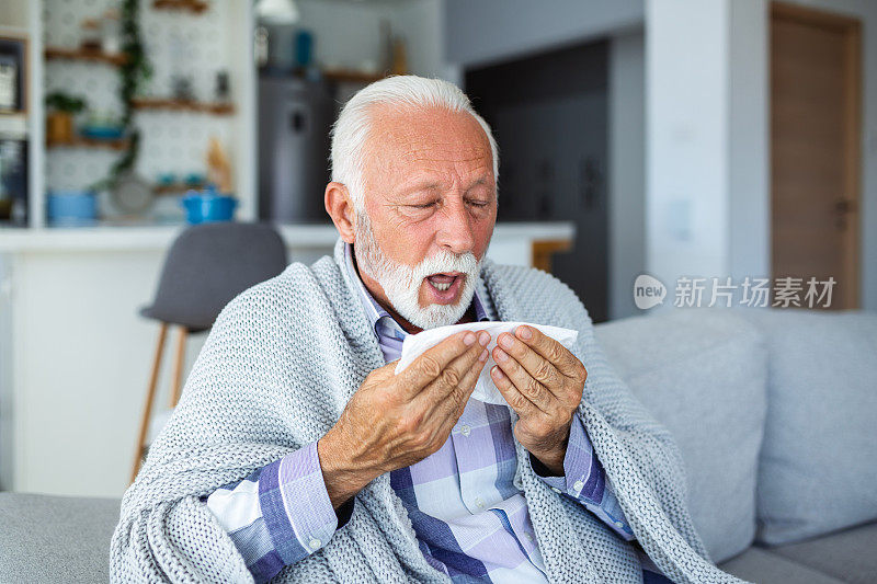 一个年长的男人，在沙发上打喷嚏，他的脸因惊讶而扭曲。手里拿着纸巾，他极力抑制声音，老人有发烧的症状，像是生病感冒季节性流感