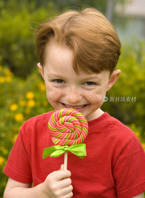 微笑红发雀斑脸男孩，孩子拿着糖果棒棒糖和花园