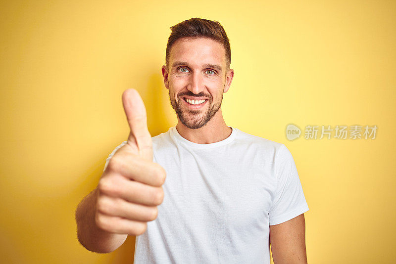 年轻英俊的男子穿着休闲白色t恤在黄色孤立的背景做快乐的拇指手势与手。赞许的表情看着镜头，表示成功。