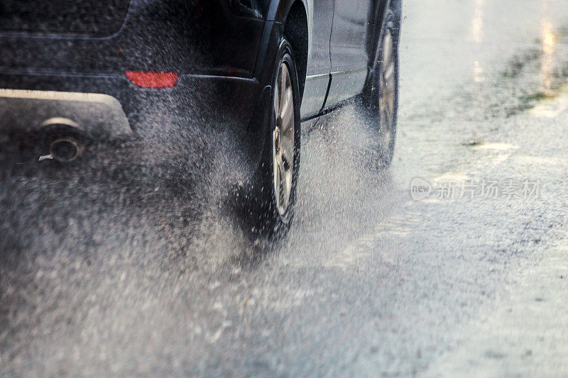 日光城市中，黑色汽车快速行驶，雨水从车轮上飞溅而出，选择性聚焦