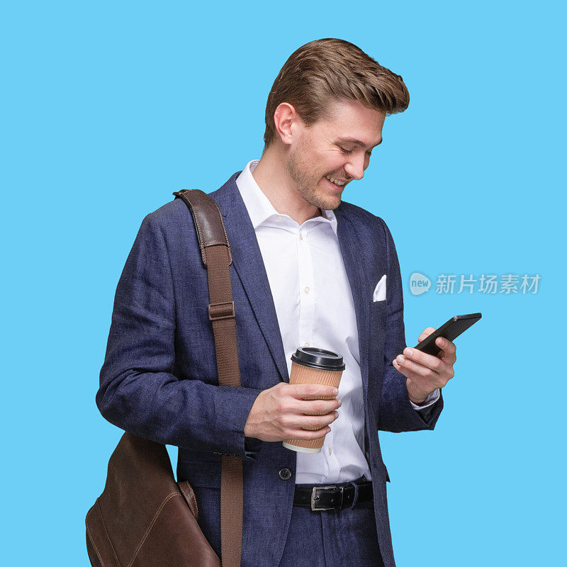白人年轻男性经理站在那里，背着小包，拿着咖啡杯，用着智能手机