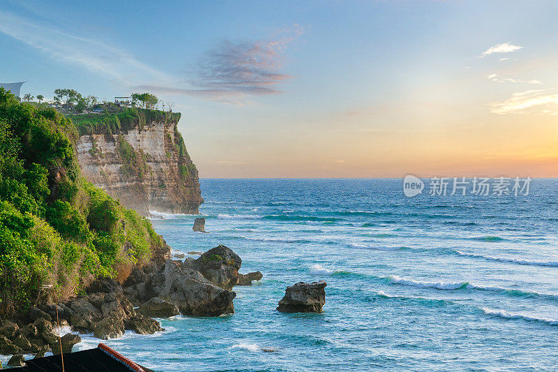 Uluwatu巴厘岛。日落在蓝色的海洋和岩石山，覆盖着热带树木。