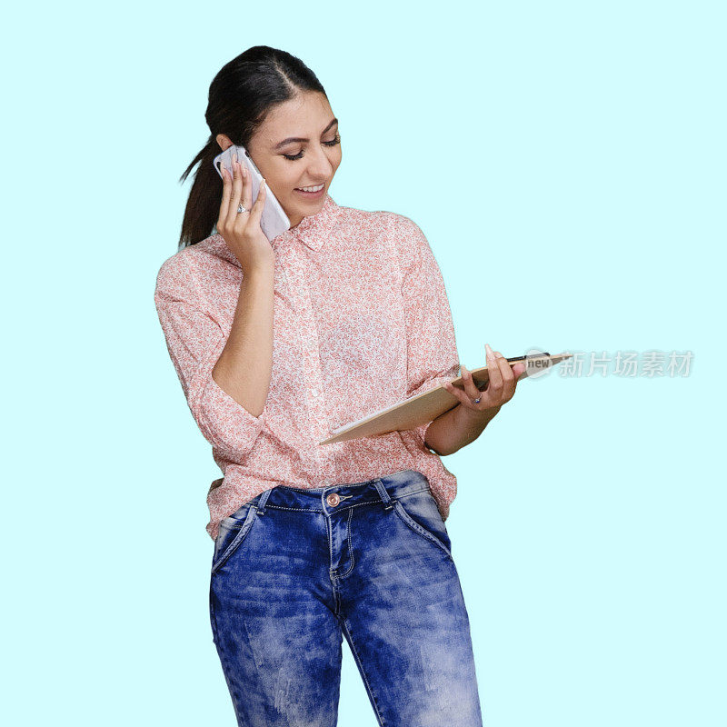 拉丁美洲和西班牙裔的年轻女性站在蓝色背景下，穿着裤子，拿着清单和使用智能手机