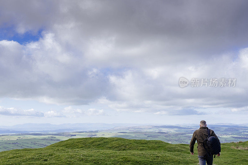 背着双肩包的男子站在山坡上，可以看到广阔的乡村景色。