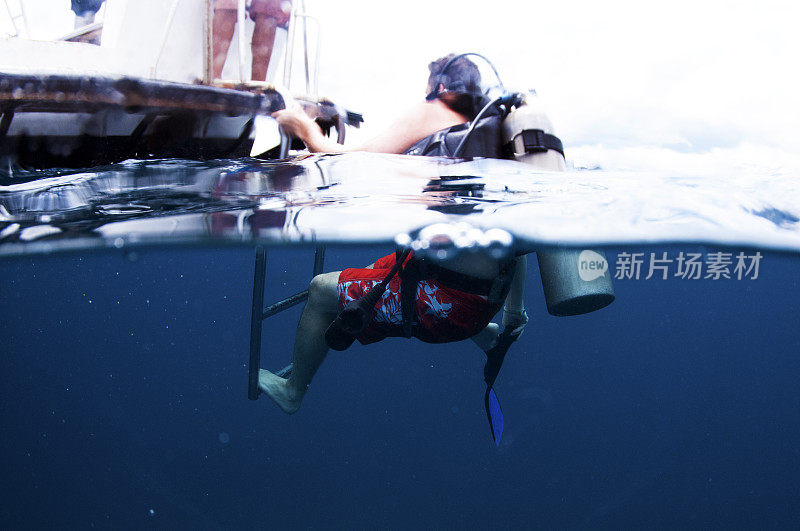 在船后面的男性潜水员取下鱼鳍回到船的水上和水下摄像机爬上梯子