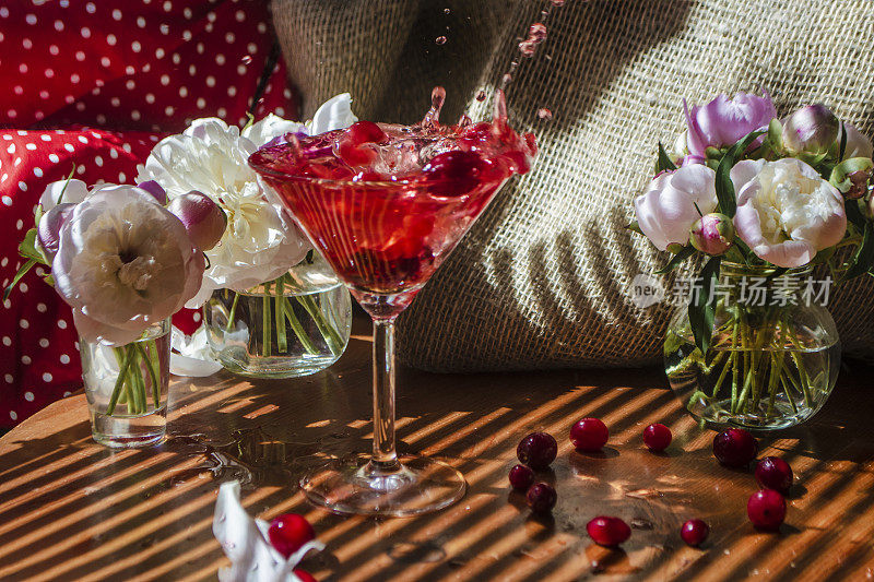 肃穆的静物，一杯红色的饮料，上面有草莓的大飞溅和掉落的浆果，被白色的花朵包围在纺织的背景上，在百叶窗的斜条纹阴影中