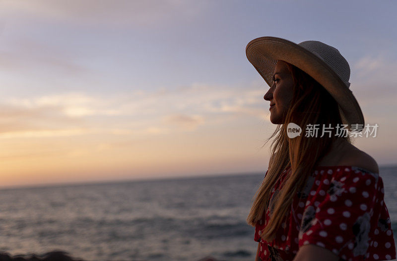 美丽的金发姑娘戴着草帽在夕阳的海上。年轻女子望着日落的天空