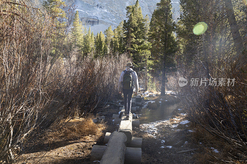 一位老人在加利福尼亚的惠特尼山上徒步旅行，他站在原木上，走在小溪上。