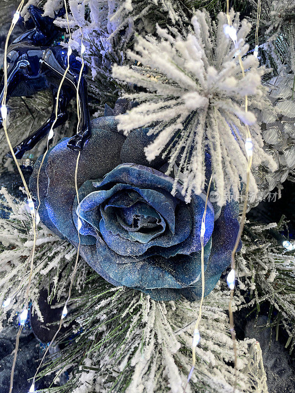形象各异的人工，蓝色的玫瑰花头装饰品挂在人工圣诞树的树枝上，云杉的针叶上覆盖着人工喷雪，背景上模糊的白色神仙灯