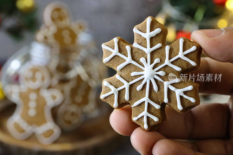 一个无法辨认的人拿着一个姜饼雪花饼干，用白色皇家糖霜装饰，姜饼人的玻璃饼干罐，迷你圣诞树，红色和金色的小装饰物，黑色背景，家庭烘焙