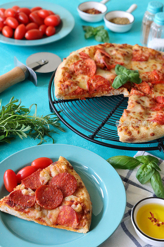 三角形的意大利辣香肠披萨放在圆形冷却架旁边的盘子里，碗里有橄榄油，方格茶巾，披萨轮，番茄，盐和胡椒瓶，蓝色背景，前景集中