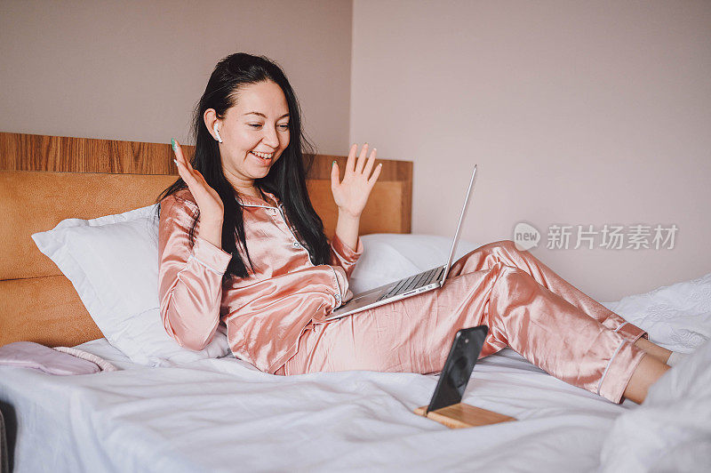 年轻快乐的黑发女子，戴着无线耳机，穿着粉色丝绸睡衣躺在床上，用智能手机和笔记本电脑，进行在线视频聊天。Blogger直播早间新闻