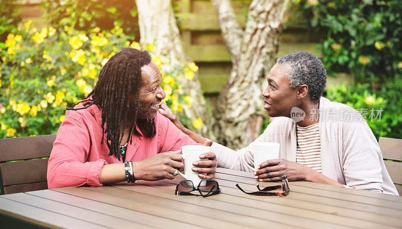 一对幸福成熟的情侣坐在室外的花园里，一起喝着咖啡。面带微笑的老男人和老女人一边喝茶休息，一边聊天