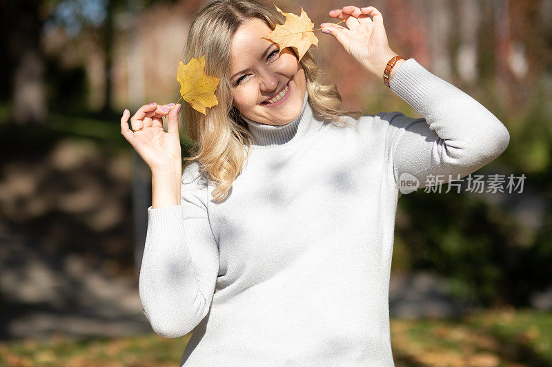 漂亮微笑的中年妇女，金色卷发，卷领毛衣，拿着黄色的枫叶近头。