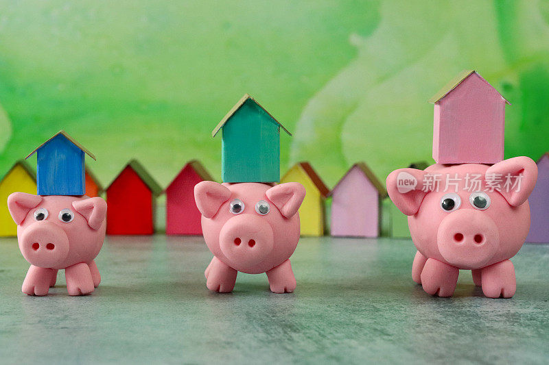 三只模型猪的形象与模型纸板房子平衡在小猪存钱罐的背部，排彩色的海滩小屋，重点在前景，斑白的绿色背景，家庭财务，房地产和假日储蓄概念
