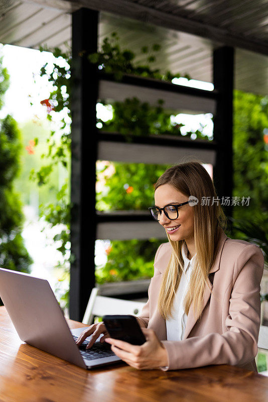 成功的商业女性坐在咖啡馆里，带着笔记本电脑，工作，写作，使用手机，桌上放着一杯咖啡。