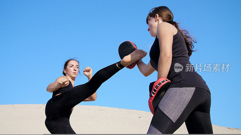 夏天，在烈日炎炎下，两个穿着黑色健身服的年轻女子在荒芜的海滩上，踢着脚，训练着打架。慢动作