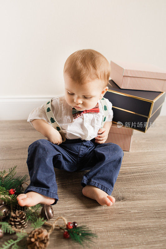 一个11个月大的圣诞男婴，穿着喜庆的红色圣诞格子格纹领结，绿色圣诞树背带，海军裤子和白色纽扣衬衫，坐在木地板上，准备迎接2022年的生日和圣诞节