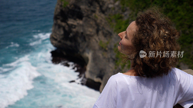 自由和幸福。一位年轻的白人女子享受着海风和微风吹过她的头发。她抬起头，迎着阳光和温暖，看着悬崖顶上的海浪。