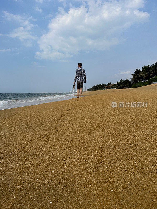 照片中，一位面目全非的男子沿着热带拉维尼亚山海滩走远，在沙滩上留下脚印，在斯里兰卡科伦坡度假，穿着短裤和方格衬衫，手拿沙滩鞋，后视图