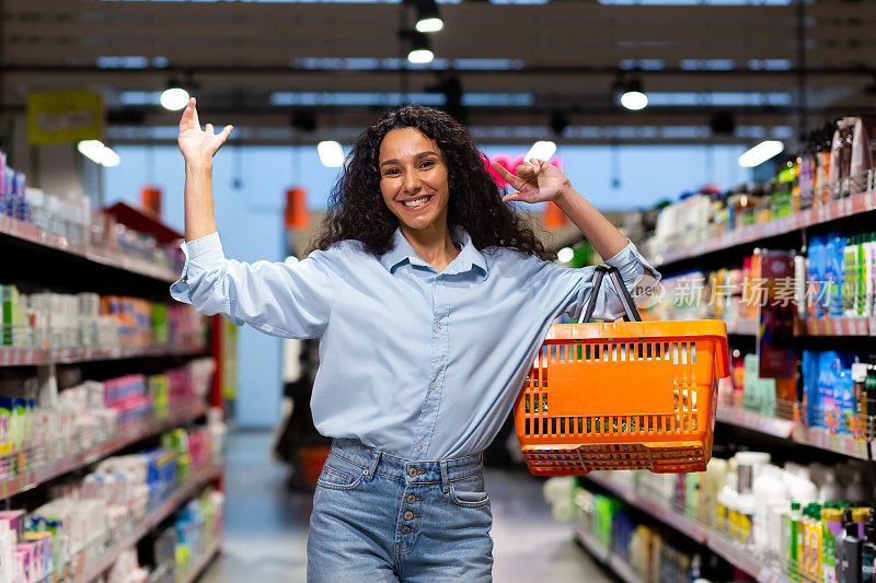 超市里一个快乐微笑的女购物者的肖像，她恐慌的女人购物，看着镜头，开心地拿着购物篮在货架上选择商品