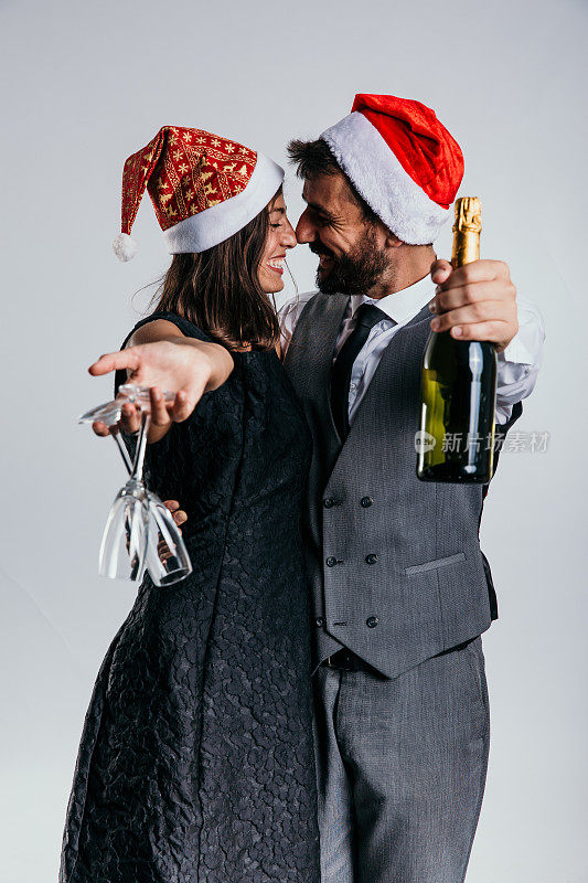 在节日的工作室背景下，一对穿着西装的夫妇在五彩纸屑和香槟的包围下戴着圣诞老人帽，为他们的庆祝活动增添了一丝异想天开。