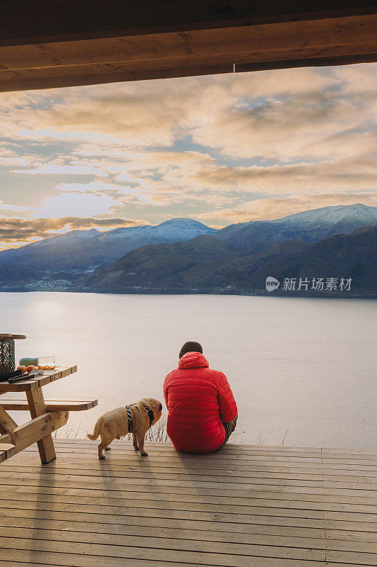 在挪威的日落时分，一名男子带着狗在露台上凝视着冬季峡湾的美景