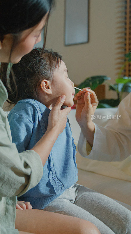 一个生病发高烧的亚洲女孩，正在家里接受医生的鼻拭子检查。生病女孩因为身体状况和家庭健康和她妈妈住在一起。垂直的屏幕。