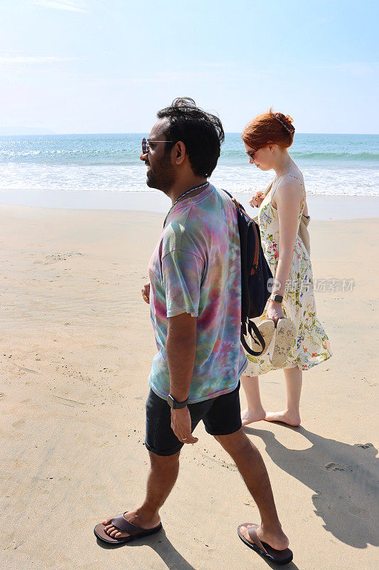 白种人，红发女人和印度男人在海边潮湿的沙滩上散步，多元文化的夫妇，晴朗的蓝天，前景的焦点