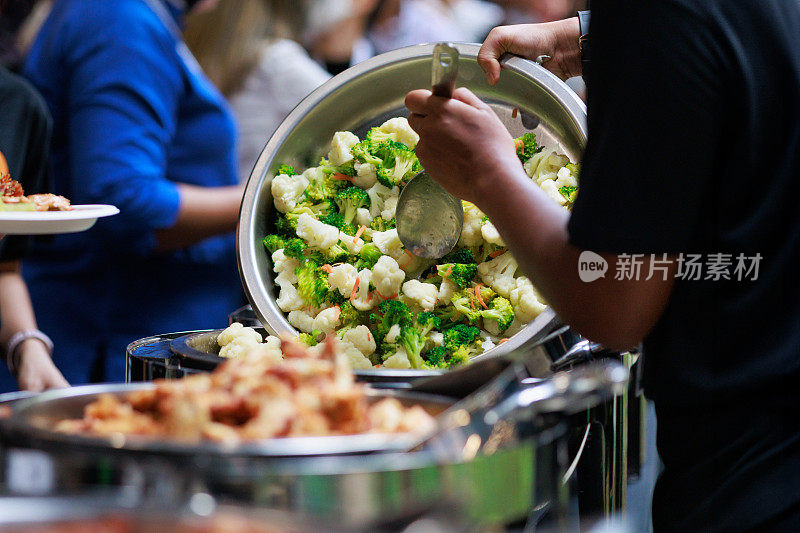 在这张裁剪过的图片中，一名厨师在自助餐队的食物托盘里重新填充新鲜煮熟的蔬菜，以确保食客们有源源不断的美味选择。