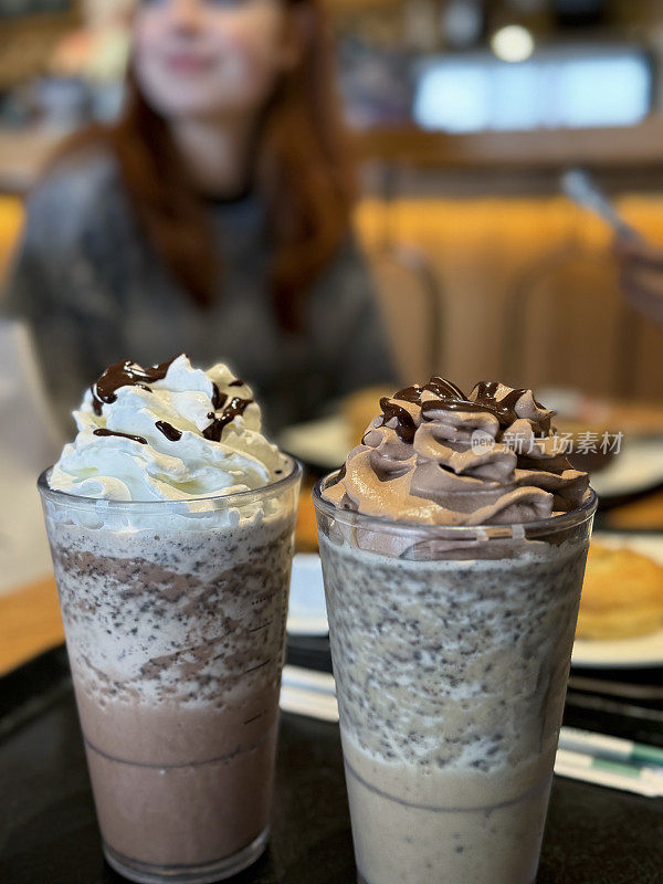 咖啡桌的特写照片，两人一起喝着摩卡星冰乐，冷饮上浇上鲜奶油或巧克力味的泡沫奶油和巧克力糖浆酱，背景是一个无法辨认的红发女人