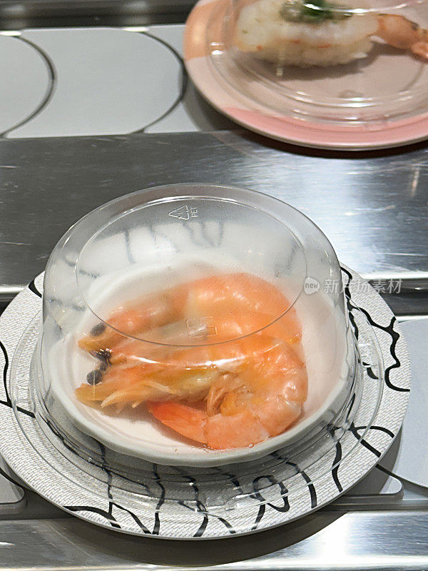 寿司店的传送带寿司(kaiten-zushi)的特写图像，有盖的盘子经过食客，甜虾(Amaebi)生鱼片，带尾虾鳕，高架视图，重点在前景