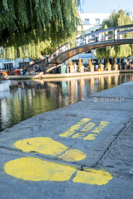 在卡姆登市场，地板上画着一个标志，要求人们在摄政运河旁边保持社交距离，背景是一座桥