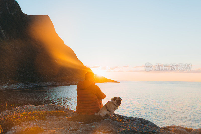 在挪威，一名男子带着他的狗在山景的海边凝视日落