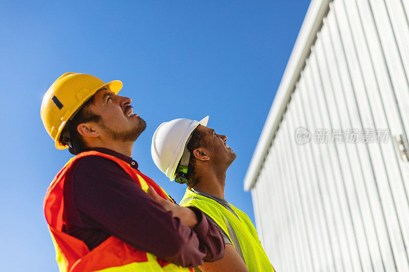 在工地上穿着PPE的黑人和西班牙裔男性建筑工人评估和讨论项目基础设施系列照片