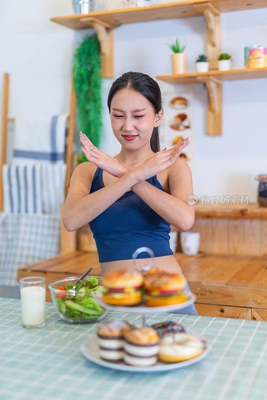 照片年轻美丽的亚洲女性女士在运动服装说不和拒绝诱惑汉堡和甜甜圈在厨房的桌子上，选择一碗沙拉与樱桃番茄和一杯牛奶的饮食和减肥健康的生活方式